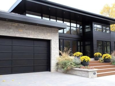 A stunning modern home entrance features sleek grey stone walls, a black garage door, and wooden stairs adorned with yellow flower pots on either side.
